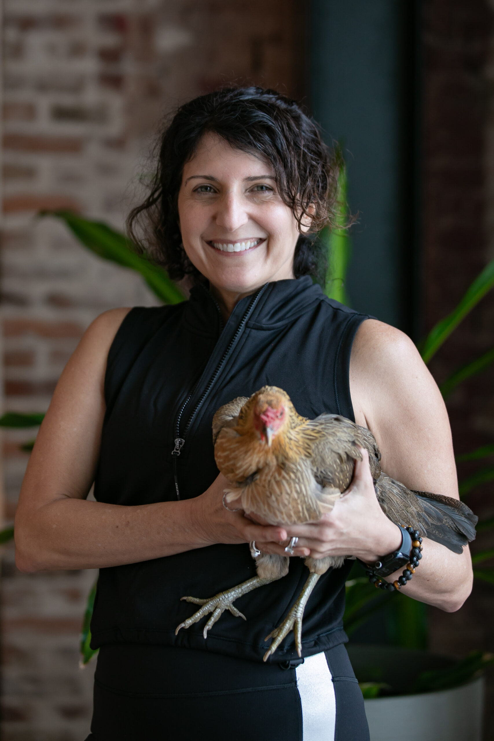 A woman smiling while holding a chicken in a casual, indoor setting with greenery and exposed brick walls, emphasizing a connection to nature.