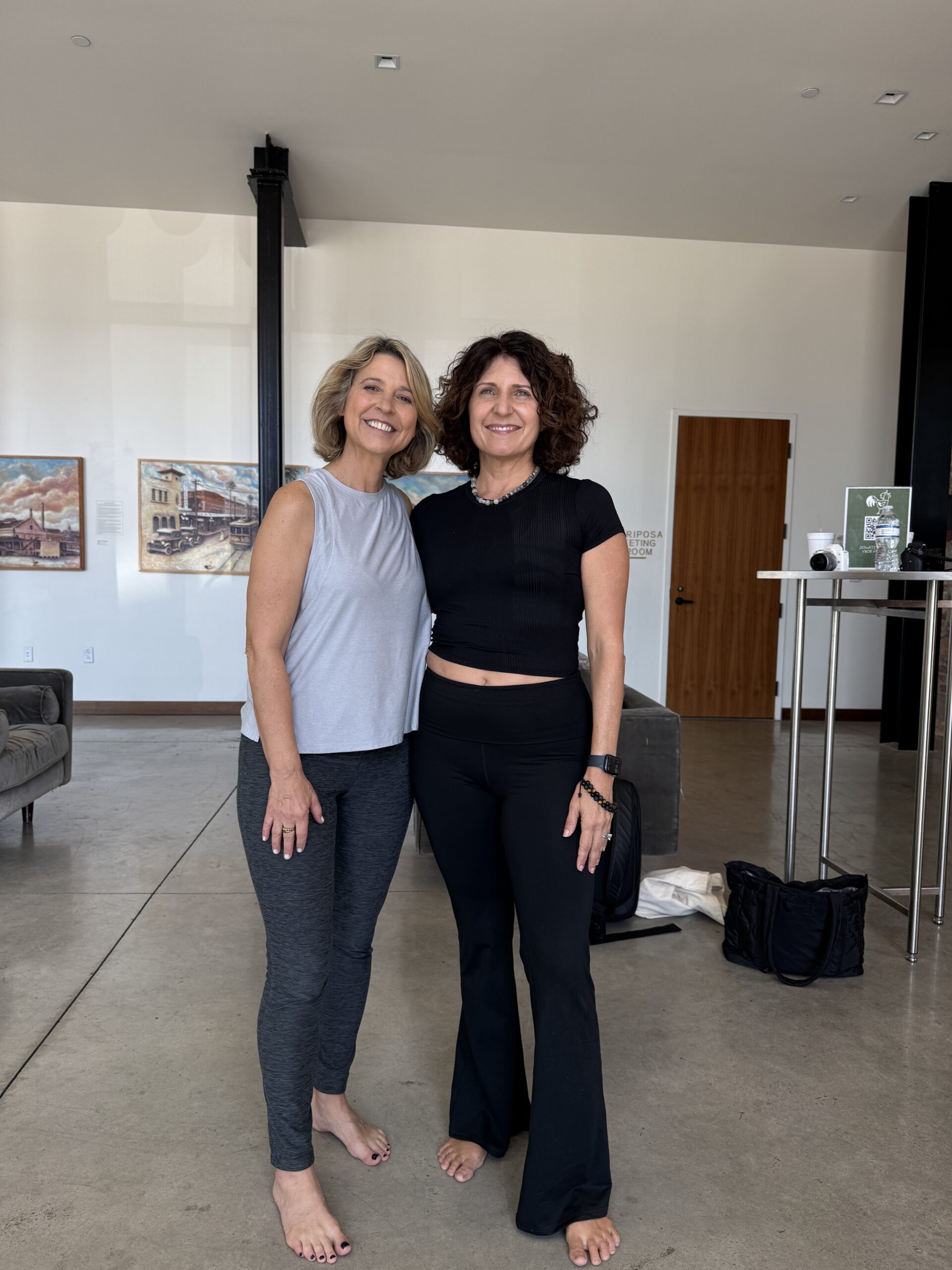 Two women pose together in a modern, bright space, wearing athletic outfits. They appear happy and relaxed, showcasing a supportive friendship.