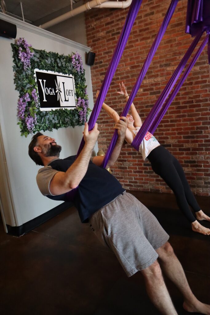 Two individuals practice aerial yoga at Yoga Loft, utilizing purple silk swings. The rustic brick wall and greenery enhance the serene atmosphere.
