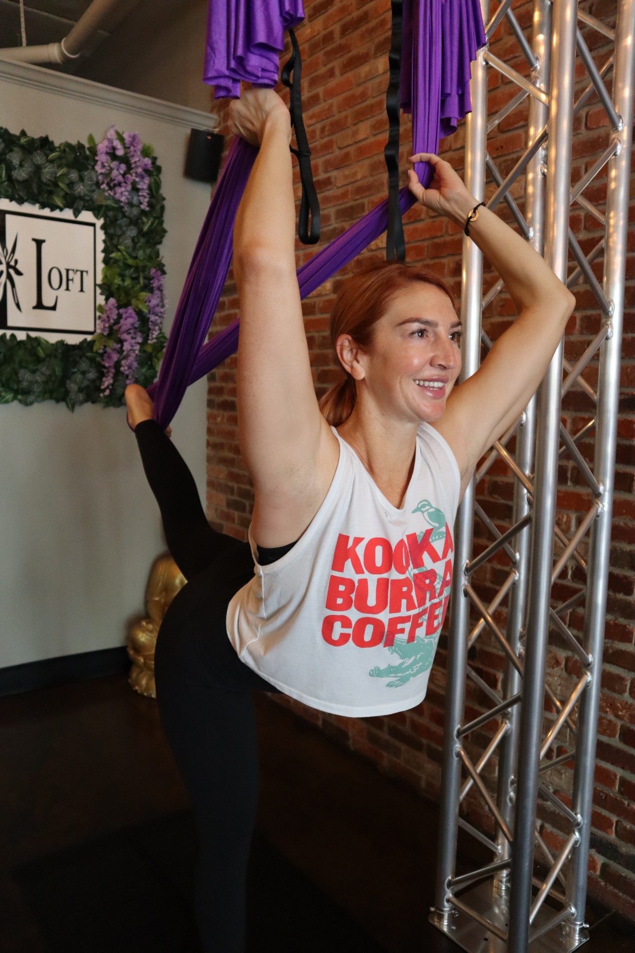 A woman performs an aerial yoga pose using purple fabric in a fitness studio with exposed brick walls. She appears focused and joyful.